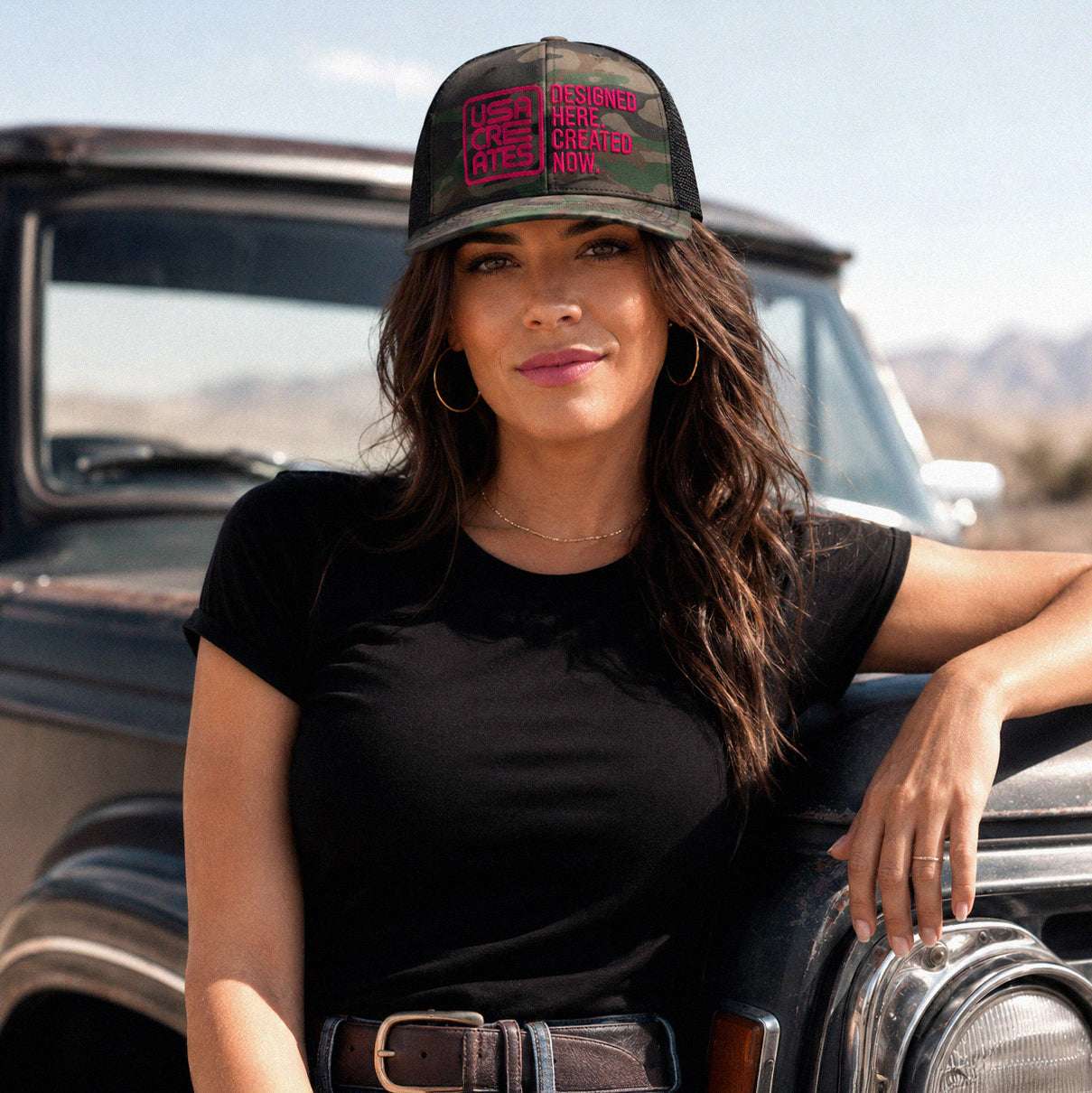 USA CREATES Camo Trucker Cherry Stitch, camo trucker hat with cherry red embroidered USA CREATES logo worn by a woman leaning on a vintage truck in an open landscape editorial portrait