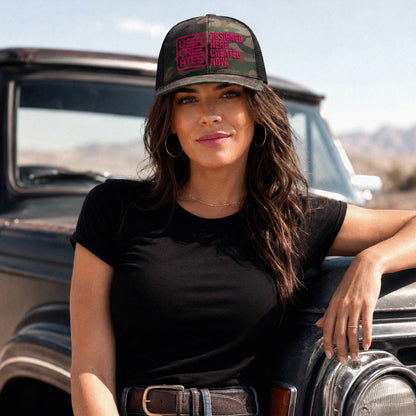 USA CREATES Camo Trucker Cherry Stitch, camo trucker hat with cherry red embroidered USA CREATES logo worn by a woman leaning on a vintage truck in an open landscape editorial portrait