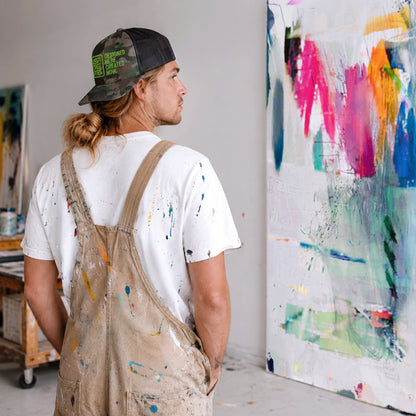 USA CREATES Camo Trucker Hat Green Stitch, shown in artist studio portrait