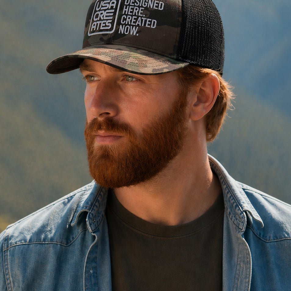 USA CREATES Camo Trucker White Stitch, camo trucker hat with white embroidered USA CREATES logo worn by a man with a beard in a mountain landscape editorial portrait